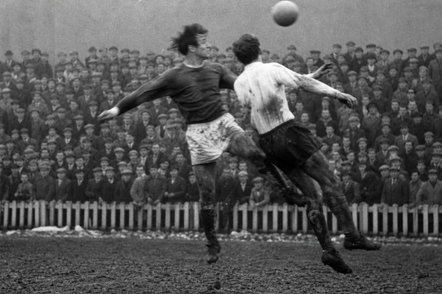 Two players challenge for a high ball on a muddy 1960s pitch in front of a terrace (picture made with AI)